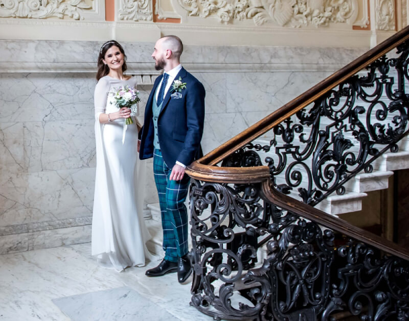 A bride and groom are captured on the stairs of a grand hall, radiating happiness in their wedding attire.