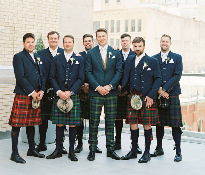 Several men dressed in colorful kilts stand together, smiling for a group photo in a scenic outdoor setting.