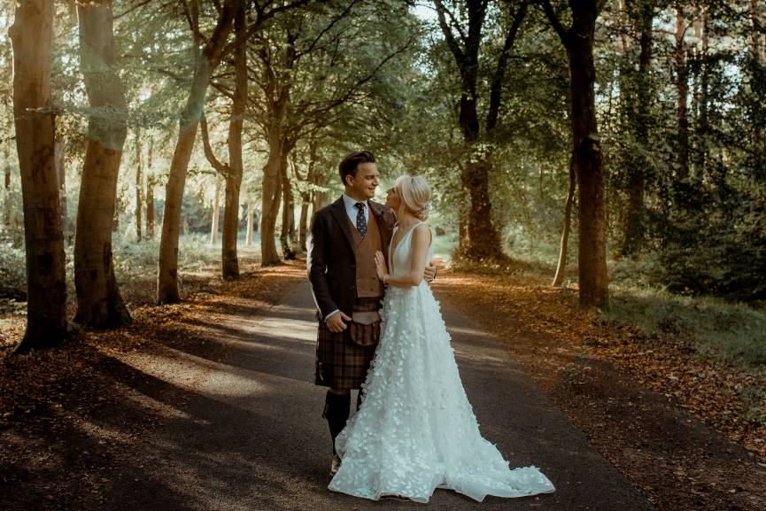 A couple in wedding attire embracing in a tranquil woodland setting, with trees and soft light creating a romantic atmosphere.