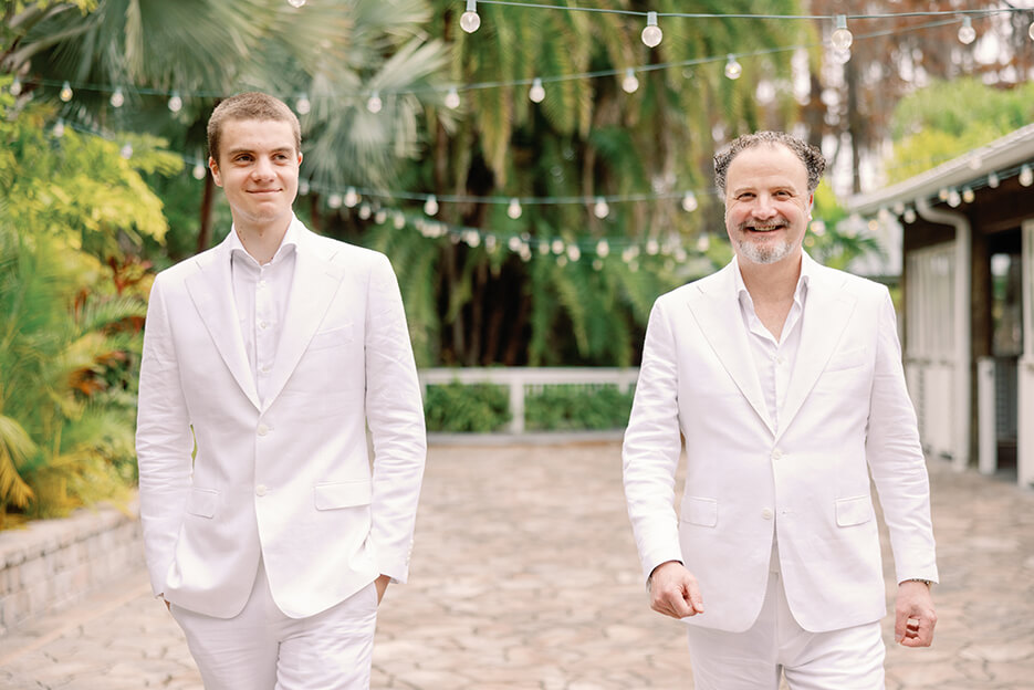 Two men dressed in white suits walk together down a picturesque path, showcasing a sense of style and friendship.