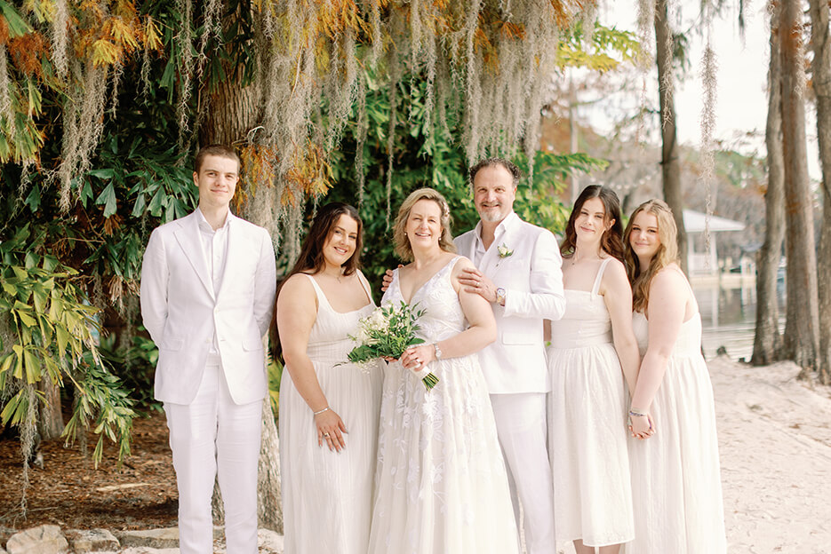 A cheerful wedding party gathers on the beach for a group photo, celebrating their special day with the ocean in the background.